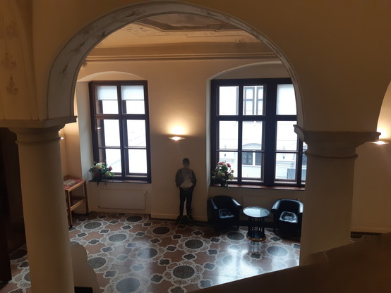 Blick von der Treppe hinunter auf den 1. Stock. Im Hintergrund 2 Fenster durch die Tageslicht scheint. Unter dem rechten Fenster 2 Sessel und 1 kleiner Tisch. In der Mitte im Hintergrund ein weiterer Aufsteller. 