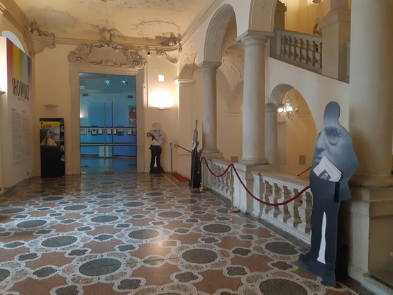 Gut ausgeleuchteter 1. Stock des Palais Lobkowitz. Rechts die Treppe in den 2. Stock mit Bögen und Säulen. Davor sowie mittig im Hintergrund weitere Aufsteller. Im Hintergrund eine blau scheinende Glastür, der Eingang zur Ausstellung "Showbiz - Made in Vienna"