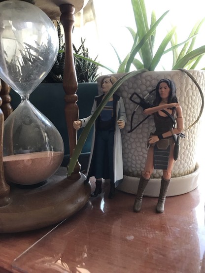 5” figures of the thirteenth Doctor and companion to the fourth Doctor, Leela beside a sand timer and surrounded by houseplants.