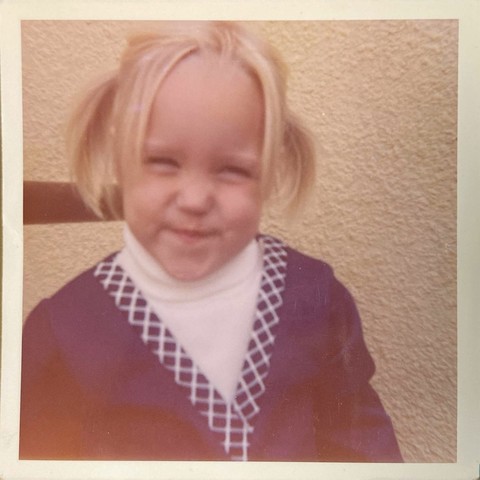 Closeup square photo of me at 3 years old. I’m standing outside in front of a cream stucco wall. I’m smiling with my mouth closed and can barely keep my eyes open because of the sun. I’m pretty certain this is from my third birthday party because I’m wearing the same outfit and my hair is the same as other photos from that day. I have straight blonde hair worn in high pigtails near the back and failed bangs because of a prominent cowlick. I’m wearing a long sleeve navy wrap dress that my mom likely made with a belt (not seen) that matches the navy and white striped edging on the dress and a white turtleneck shirt underneath. Also not seen: white knee socks and red Mary Jane shoes.