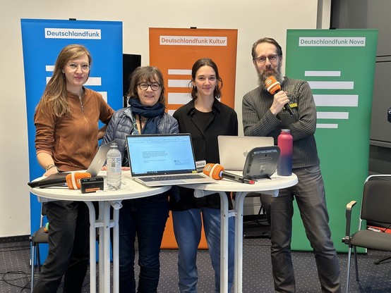 Jenny Genzmer, Vera Linß, Pia Behme & Marcus Richter an einem Tisch auf dem Computer stehen. Im Hintergrund Aufsteller von Deutschlandfunk (blau), Deutschlandfunk Kultur (orange) und Deitschlandfunk Nova (grün)