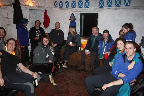 A group of about 20 people and 2 dogs, in a small room with concrete floor and whitewashed stone walls. Saltire flag bunting and waterproof jackets hanging up. 