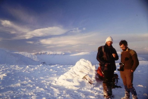 Two people at a cold summit cairn. Snow covered ground and blue sky with clouds.