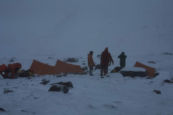 A group of small tents in the snow with some rocks showing through. A few people stood around wrapped up against the cold. Dull day.