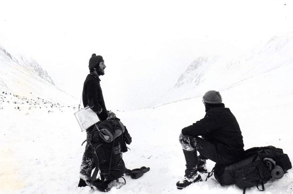Black & white pic, two people wrapped up against the cold, one sat one stood, with large rucksacks, looking at the view. Snowy, with crags visible in the mist in the distance.