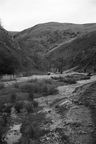 A black-and-white shot of Dovedale in the Peak District, UK. The river Dove cuts through a winding valley with more hills in the distance; it's burst its bank and the usually walkable tracks either side of the river are flooded. There's also been some small landslides, making the valley pretty tough to cross.