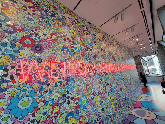 Long gallery wall at The Asian Art Museum of San Francisco, covered in brightly-colored cartoon daisies popularized by Takashi Murakami. A red neon sign in block letters runs the length of the wall, "Weirdly Lovable, Lovably Weird". Sunlight illuminates the wall from exterior windows. 
