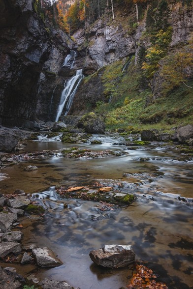 Se puede ver una cascada al fondo hacía la izquierda, con algo de bosque en la derecha y rocas y agua en primer plano.