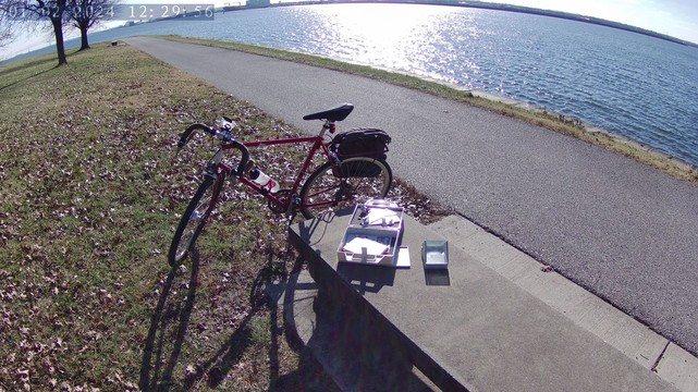 A picture of a bicycle parked near the water's edge.  Adjacent to it is a stone bench on which rest an artificial horizon and an open box containing a Davis Mark 3 marine sextant.