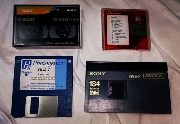 4 old format s laid out : Basf Hi-8 video cassette. A blank red minidisc with typed label containing music by Saint-Sean's, a blue 3.5" floppy disc install disc for Photogenics, and a grey Sony DVCam video cassette, full sized 184 minutes.