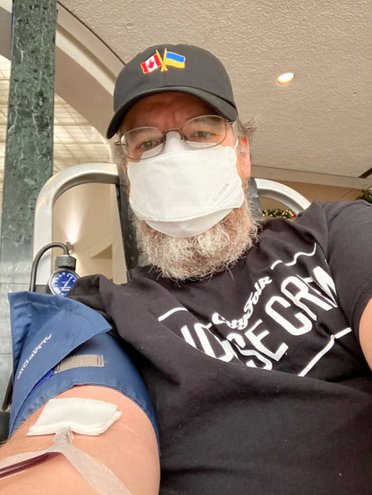 Selfie of a white guy with glasses, a grey beard, and a ball cap, wearing a mask, in process of giving blood.