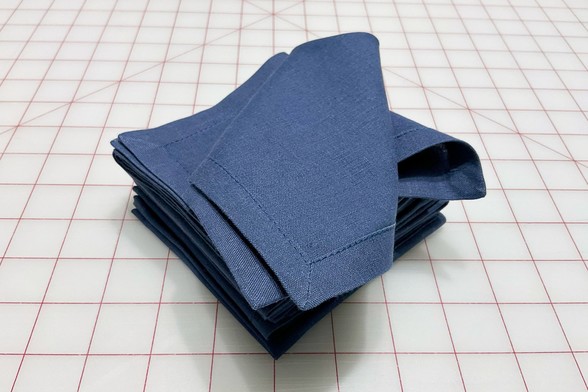At stack of freshly sewn 9-inch reusable cotton/linen napkins with mitred corners and half-inch hems sit atop a large cutting mat. The top napkin shows the underside of the napkins.