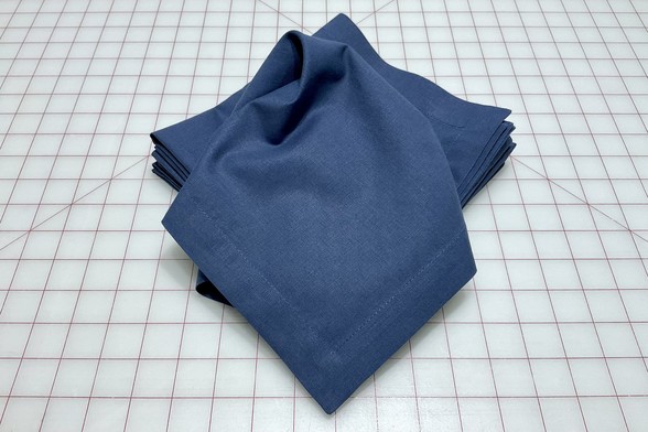 At stack of freshly sewn 16-inch reusable cotton/linen napkins with mitred corners and one-inch hems sit atop a large cutting mat.