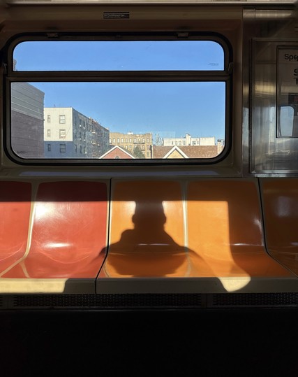 Shadow on subway seats. The seats are orange and yellow. Out the window is a blue sky and Bronx apartment buildings. 