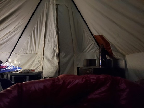a serene scene of the inside of canvas hot tent. the foreground has a down quilt. the background shows a wood stove.