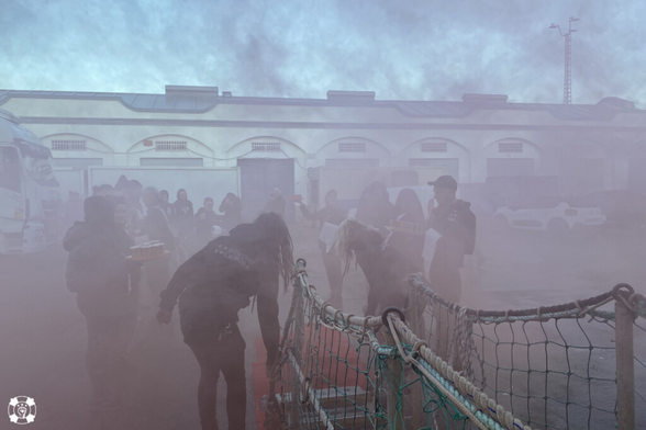 Blick von der SEA EYE 4 auf Kai und Lagerhalle in Burriana. Viele Menschen warten dort eingehüllt in Rauch. Ein roter Teppich ist ausgerollt.