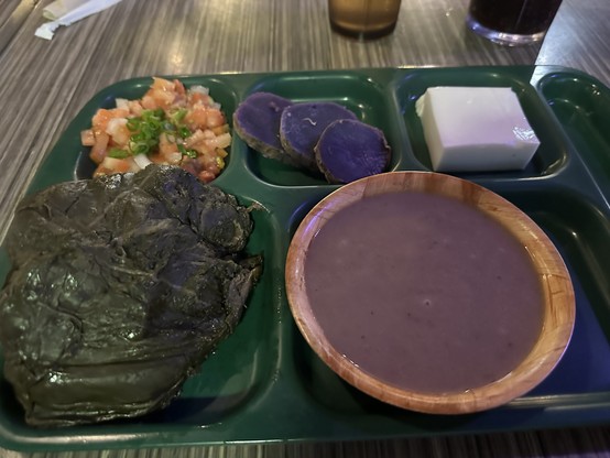 A tray of food including a green lau lau, purple ʻuaʻa or sweet potato slices, a white block of haupia, a lomi lomi salmon, and a bowl of poi. A diet coke is visible in the background. 