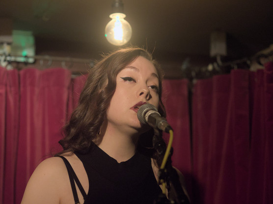 Head and shoulder of Scarlett Fagan singing into a microphone on a stand. A glass lamp hangs above her providing little illumination. In the background the purple velvet curtains of the Green Note basement bar.