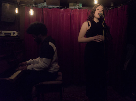 Scarlett Fagan singing into a microphone on a stand. She is wearing a black dress with her hands clasped against her chest. To her right a pianist is playing an upright piano. The scene is dark and underexposed due to the only lighting being a few small lamps hanging above the musicians and my wide lens being too slow.