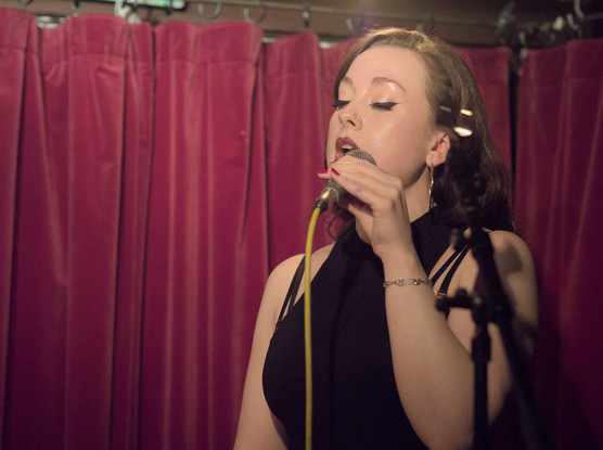 Scarlett Fagan holding a microphone to her mouth.  She is wearing a black dress with her shoulders bare. Her eyes are closed as she feels her music.