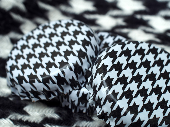 an indoor photograph close-up of three metal bottlecaps. the bottlecaps are metal and are painted with a black and white houndstooth pattern. the bottlecaps rest on a woven fabric scarf that is actually woven with a black and white houndstooth pattern. there are no people, animals, or vehicles visible in this picture.