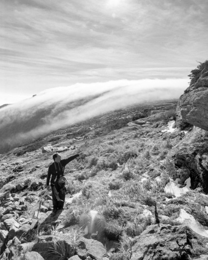 Black and white photo of a man standing on a mountain slope, facing and pointing with one arm, uphill. Behind him, a stream of clouds flows over the ridge of the mountain down into the valley.