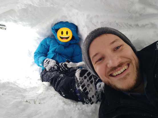 A man with facial hair, wearing a grey beanie smiling while a young child sits behind him wearing a blue jacket, snow pants, and gloves while holding a small shovel. The child's face is covered by a smiling emoji. They are both inside and under a small snow fort.