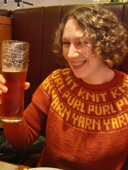 A woman with short curly hair and wearing a red-brown round yoke knit sweater, with the words “knit”, “purl”, and “yarn” in yellow yarn knit into the yoke, is holding up a beer and smiling into the camera