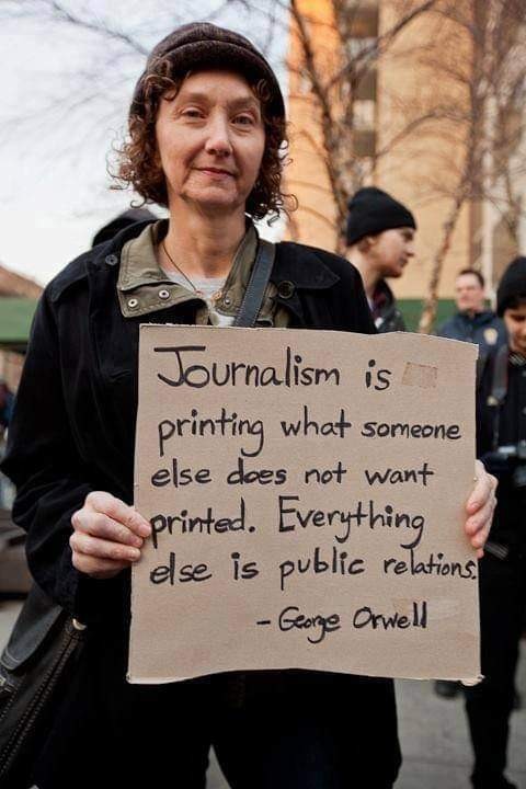 Alt-text: A woman holding a hand-written cardboard sign that says: Journalism is printing what someone else does not want printed. Everything else is public relations. —George Orwell
