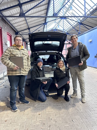 In einer Gewerbehalle des Kulturhauses Alter Schlachthof mit Glasdach auf Stahlträger steht ein dunkler Pkw mit offenem Kofferraum. Im Kofferraum liegen gestapelte Laptops. Vor dem Kofferraum stehen 4 Personen. Ein Foto zeigt Dominik Lux und Anna Ulbrich (Ferber-Software), Michael Osterhof (Geschäftsführer Alter Schlachthof) und Thomas Schirdewahn (Ihr Soester Computer-Assistent)