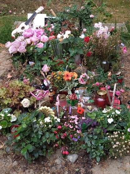 Grabschmuck am Grab der Künstlerin und Musikerin Francoise Cactus. Zwischen vielen Blumen und einem Kaktus (!) finden sich unzählige kleinere und größere Accessoires, die nicht auf einmal zu erfassen sind.
