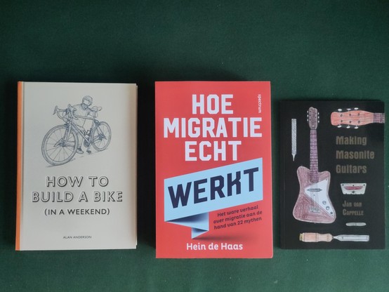Three books, from left to right: "how to build a bike (in a weekend)" by Alan Anderson - light grey front, man holding a bike, title and author below; "how migratie echt werkt" by Hein de Haas - red cover, title in white, except the word 'werkt', which is in black, in a light blue box, author's name in white below; "making Masonite guitars" by Jan van Capelle - black cover, with a guitar, guitar neck and some tools, title and author in brown.