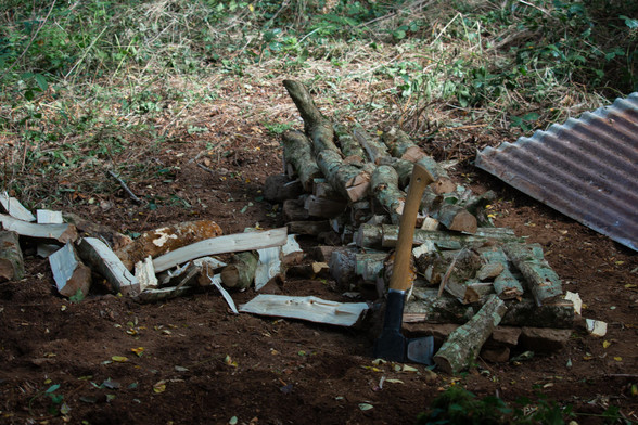 Début d'un tas de bois de chauffage en forêt avec une hache à fendre posée contre lui. D'un côté des bûches tout juste fendues et de l'autre une tôle un peu rouillée.