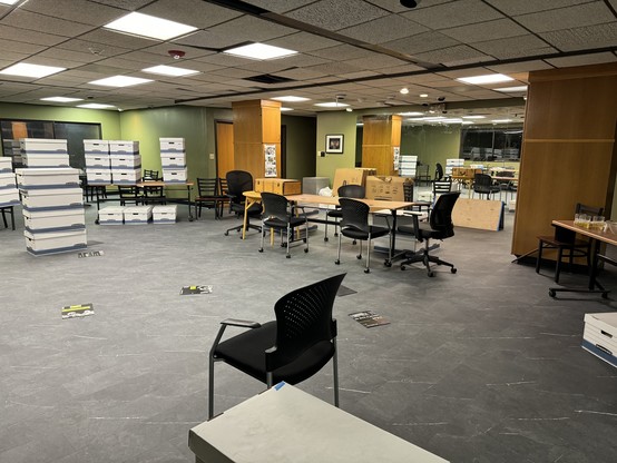 Rehearsal room with desks, chairs, and piles of boxes. There are papers scattered on the floor and the space appears disorganized.