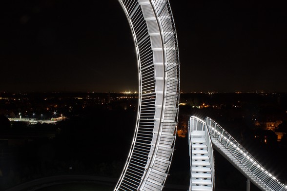 Fußgängerachterbahn Tiger & Turtle, nachts angestrahlt in hellem weißen Licht im Hintergrund leuchtende Industrie und Stadtkulisse