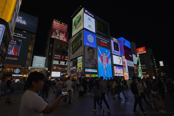A street scene at night, in the background building clad entirely in giant video screen advertisements, in the mid ground, a stream of people walking left to right of frame and back. In the foreground, a young man viewed in profile has a glass sphere the size of an orange, resting on his outstretched palm. The entire scene is contained and flipped within it.