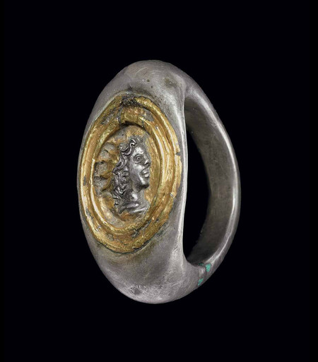 Roman silver finger ring with the head of Sol-Helios on a golden background. The solid hoop flat on the interior, rounded on the exterior, expanding to the broad angular shoulders, the bezel centered by a relief radiate bust of Helios in profile to the right, framed by raised bands, the bezel overlaid in sheet gold, the face and hair of Helios cut out to reveal the silver below.