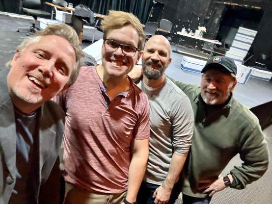Group selfie of the cast and director of The Lehman Trilogy at Playhouse on the Square with a stage and theater equipment in the background.