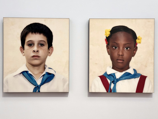 These are two portrait photographs by Desiree Dolron. They are part of a reportage on Cuba. On the left photograph is a white schoolboy, and on the right photograph is a black schoolgirl. They both wear white dress shirts with a blue scarf. The boy has black hair and blue eyes. The girl wears yellow and red hair clips, and the red straps of her school dress are visible as well.