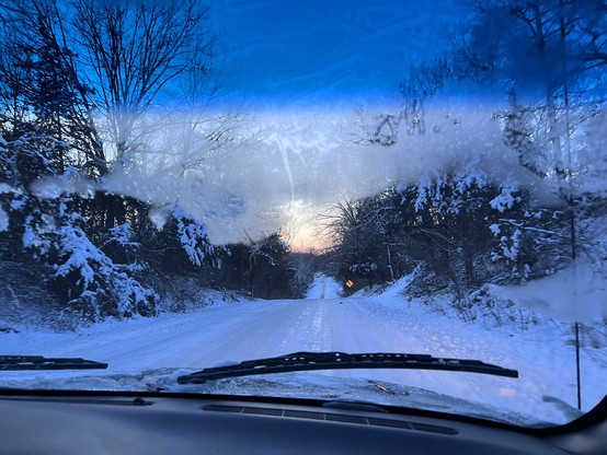 Driving down a snowy road with frosted trees on either side and a pink sky of sunset directly ahead. The windshield on my passenger side still holds frost. 