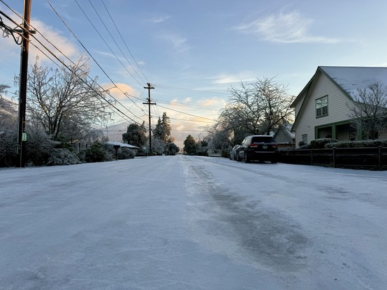 A residential street covered in snow during late afternoon with houses and parked cars on the side, power lines overhead, and a sunrise in the background.
