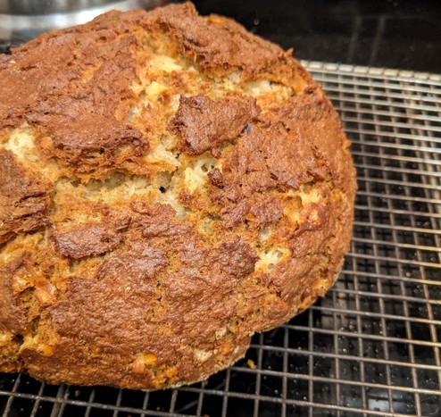 A round, crusty loaf of bread on a metal grid cooling rack. The bread is rustic with cracks and crags of cheddar cheese and I may not wait for it to cool before eating some.