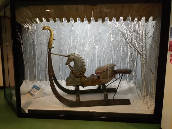 A museum case containing a one or two person sleigh.  The runners curve up in front to forma swan's head, the front seat is padded and has a unicorn's neck and head.  There is a small pillion seat behind.  The sleigh is guilded in places, the unnicorn' part is whilte.  The case floor is white like snow, the back painted a pale grey, the colour of a winter sky, and bare white braches stand like small trees in front of it.  There is a museum interpretation label angled away from the camera.