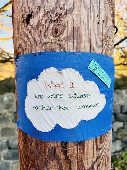 An embroidered cloth attached to a telegraphy pole. The stitching says: what if… we were citizens rather than consumers. #LeedsDreams for our future