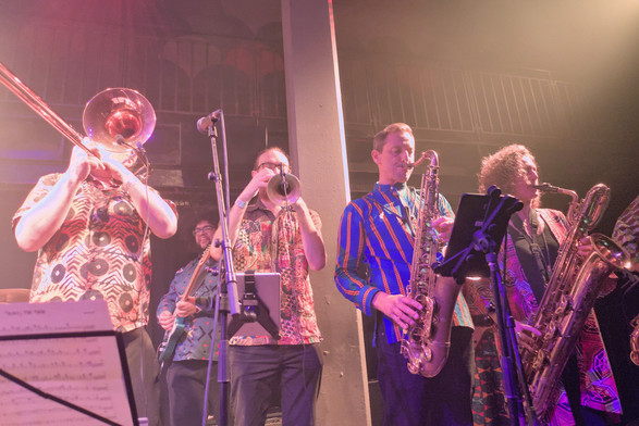 The horns section. 3 men are playing trombone, trumpet, and saxophone, and a women is playing another sax. A guitarist is in the background partially obscured behind the trumpet player.