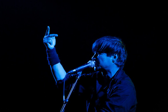Frank Z., der verstorbene Sänger der Band Abwärts bei einem Live-Auftritt in blauem Licht. Er steht vor einem Mikrofon und hebt die Hand. 

Bildquelle: Wikipedia -> https://commons.m.wikimedia.org/wiki/File:Abw%C3%A4rts2009FZ.jpg#mw-jump-to-license