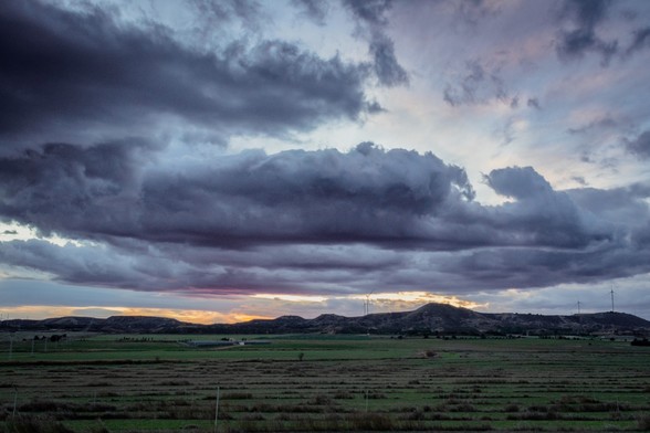 Se puede ver un paisaje donde más de la mitad está ocupado por grandes nubes que parecen de tormenta con el atardecer de fondo. Bajo ellas, campos verdes.

You can see a landscape in which more of it are stormy big clouds at sunset. Below the clouds, you can see green fields.