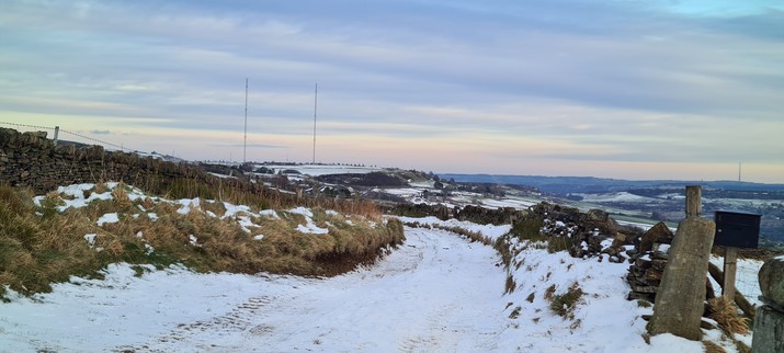 A snow covered country road disappears around a bend. In the distance along the same line, there are two 150m tall antennae on a hilltop. 