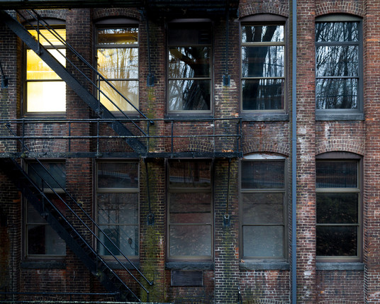 Color photo of the side of a brick building, cropped to show 10 tall windows and part of a fire escape. Two of the windows are lit from within and the rest are not. 