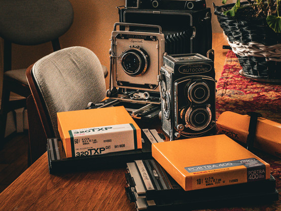 Color photo of photography equipment and film on a table. There are several 4x5 film holders and boxes of film, a Minolta Autocord twin lens reflex camera, and a Speed Graphic 4x5 press camera. An effect has been applied to the image to give a nostalgic feel.